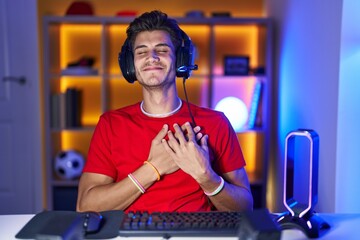 Young hispanic man playing video games smiling with hands on chest with closed eyes and grateful...