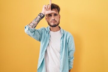 Young hispanic man with tattoos standing over yellow background making fun of people with fingers...