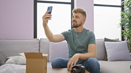 A young bearded man takes a selfie in a modern living room with a box and dslr camera.