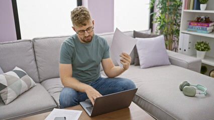 Handsome caucasian man with blue eyes and a beard reviews documents on a laptop in a modern living...