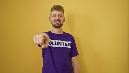 A handsome young man with blue eyes and beard wearing a purple 'volunteer' t-shirt points and...