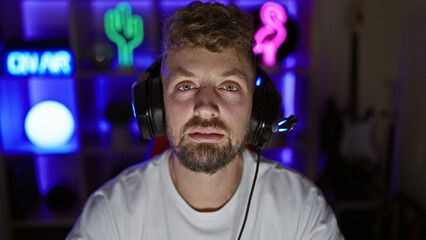 Caucasian man with blue eyes and beard wearing headphones in a dark gaming room at night