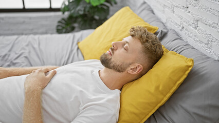 Young bearded man sleeping peacefully in a modern bedroom with yellow pillows and gray bedding.