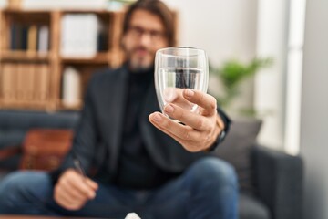 Middle age man psychologist holding glass of water sitting on sofa at psychology clinic
