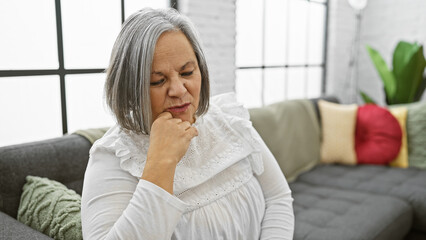 A pensive, grey-haired woman sits thoughtfully in a cozy, modern living room, reflecting on mature adulthood.