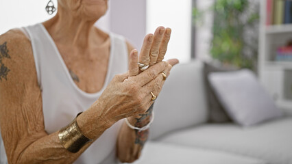 Senior grey-haired woman suffering intense hand pain, sitting in living room sofa at home, elderly...