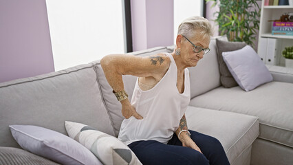 Unhappy grey-haired senior woman suffers severe backache, sitting on home sofa. worried, mature female adults grapple with serious spine problem indoors in middle age.