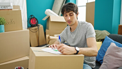 Young hispanic man writing on document sitting on sofa at new home