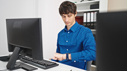 Young hispanic man business worker using computer looking watch with serious face at the office