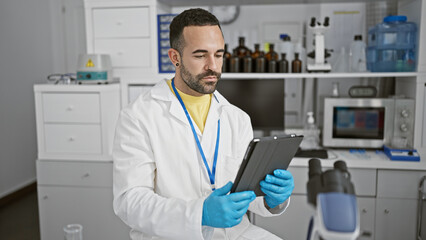 Naklejka na ściany i meble A young hispanic man with a beard wearing a lab coat and gloves holds a tablet in a medical laboratory setting.