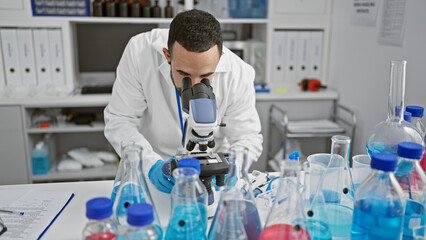 Hispanic man in lab, using microscope, surrounded by scientific equipment.