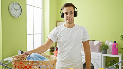 A young man in headphones holding a laundry basket stands in a brightly lit home laundry room.