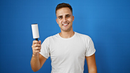 Smiling young hispanic man holding a lint roller against an isolated blue background