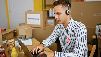 Handsome young man with headset working on laptop in a warehouse office surrounded by boxes and food supplies.