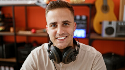 Smiling young man with headphones in music studio surrounded by musical instruments and audio...