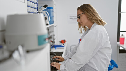 Engrossed young blonde scientist woman meticulously working on research through computer in bustling laboratory