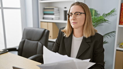 Attractive young blonde female worker deeply concentrated, thinking about business-related documents at her workplace, depicting strength and elegance.