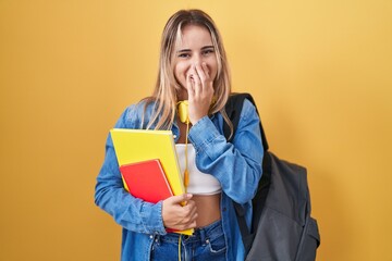 Young blonde woman wearing student backpack and holding books laughing and embarrassed giggle...