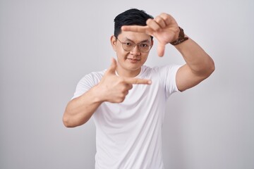 Naklejka premium Young asian man standing over white background smiling making frame with hands and fingers with happy face. creativity and photography concept.