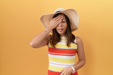 Middle age chinese woman wearing summer hat over yellow background yawning tired covering half face, eye and mouth with hand. face hurts in pain.