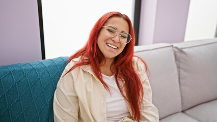 Cheerful young redhead woman, comfortably wearing glasses and resting on a cozy sofa, radiates joy and confidence, as she smiles positively in the indoor comfort of her warm living room.