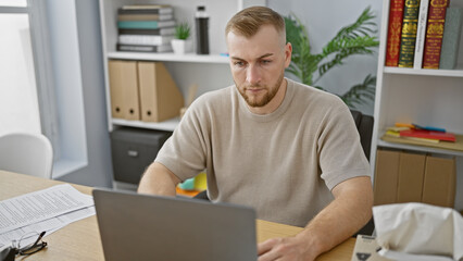 Bearded young man working on laptop in modern office with plant decoration.