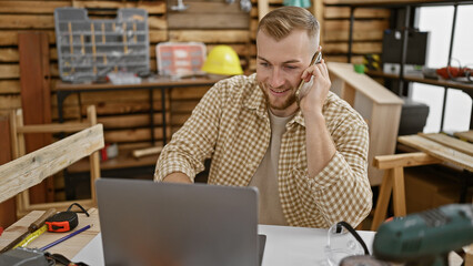 A young caucasian man with a beard works on his laptop in a carpentry workshop while talking on the phone.