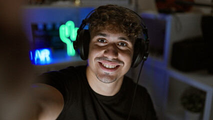 Young, attractive hispanic guy, a passionate streamer, making selfie memories! smiling for the camera from the heart of his gaming room, creating an entertaining stream. © Krakenimages.com