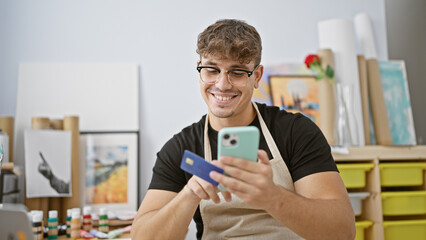 Ecstatic young hispanic man artist brimming with joy as he uses his credit card on a smartphone for...