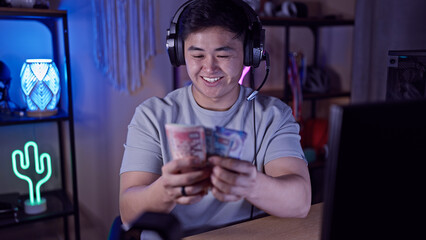 A young asian man smiles while holding hong kong dollars in a gaming room at night.