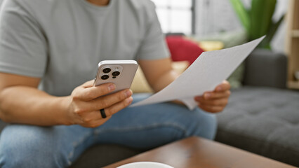 A young asian man checks a document while holding a smartphone in a modern living room setting.