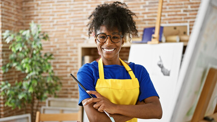 African american woman artist standing with arms crossed gesture holding paintbrushes at art studio