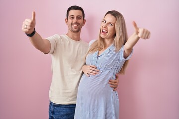 Young couple expecting a baby standing over pink background approving doing positive gesture with hand, thumbs up smiling and happy for success. winner gesture.