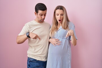 Young couple expecting a baby standing over pink background pointing down with fingers showing...