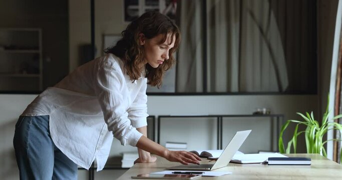 Focused young small business owner woman using laptop in modern office, standing at large workplace table, typing on computer, looking away, thinking, working on startup