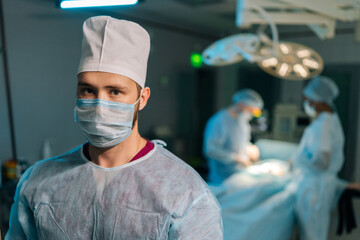 Close-up portrait of male doctor in surgical uniforms and masks standing posing looking at camera in operating room. Diverse professional team of surgeons performing operation on background