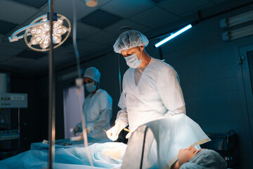 Portrait of male surgeon and African female nurse performing operation under electric lamp in dark...