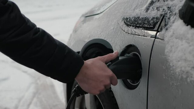 Male hands unplug power connector from EV, snow closeup. Unrecognizable person disconnects charging cable from an electric car. Charging batteries an electric vehicle or PHEV in winter