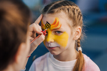 A woman professional artist paints with a brush, a colored pencil on the face of a child, a beautiful girl, face painting, makeup, watercolor drawing at a party. Photography, creative process.
