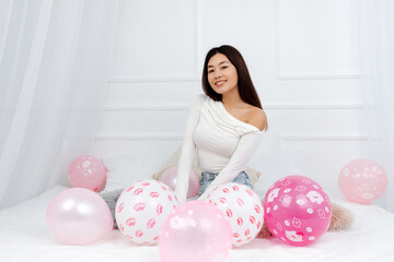 Cute brunette asian woman sitting around pink balloons and looking at camera