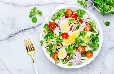 Fresh salad with squid, eggs, cherry tomatoes, onion, lamb lettuce and olive oil with lemon juice dressing, marble table background, top view