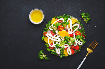 Fresh salad with squid, oranges, cherry tomatoes, lamb lettuce and olive oil with lemon juice dressing, black table background, top view