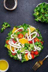 Fresh salad with squid, oranges, cherry tomatoes, lamb lettuce and olive oil with lemon juice dressing, black table background, top view