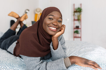 Portrait of young trendy african woman in muslim headscarf smiling at camera while lying on bed - obrazy, fototapety, plakaty