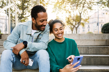 Two young latin american people using smart phone together sitting outdoors. Cheerful african...