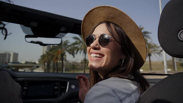 Cheerful young adult Latina co-driver enjoying sunny day in smiling convertible car. Attractive millennial woman with hat and glasses on tourist road trip on summer vacation. People in leisure time