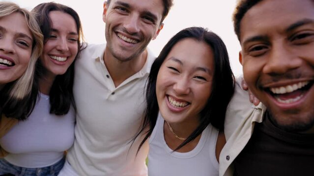 Multirracial group of young smiling friends looking at camera in the park hugging in community. Happy millennial people laughing taking selfie having fun together outside. Cheerful university students