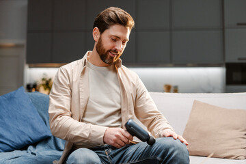 Portrait of attractive handsome bearded man using massage gun, doing self massage, massaging knee 