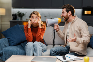 Angry man holding megaphone, shouting loudly to his girlfriend, gesturing while sitting on sofa