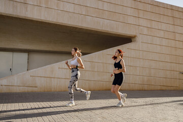 Obraz premium Two young women in sportswear are running on modern buildings background. Active lifestyle concept